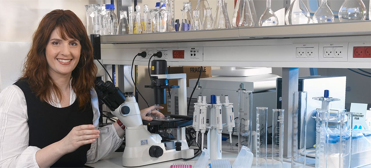 Female scientist smiling at a microscope in a laboratory, surrounded by glassware, pipettes, and lab equipment, highlighting her role in scientific research and experimentation.