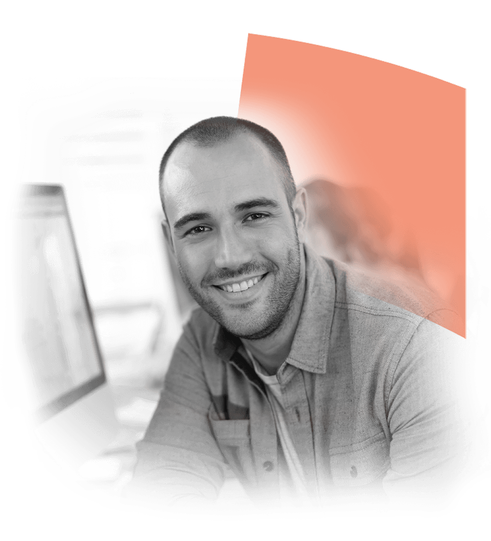 Smiling man sitting at a desk in an office environment, wearing a casual shirt, with computer screens in the background, conveying a professional and approachable atmosphere.