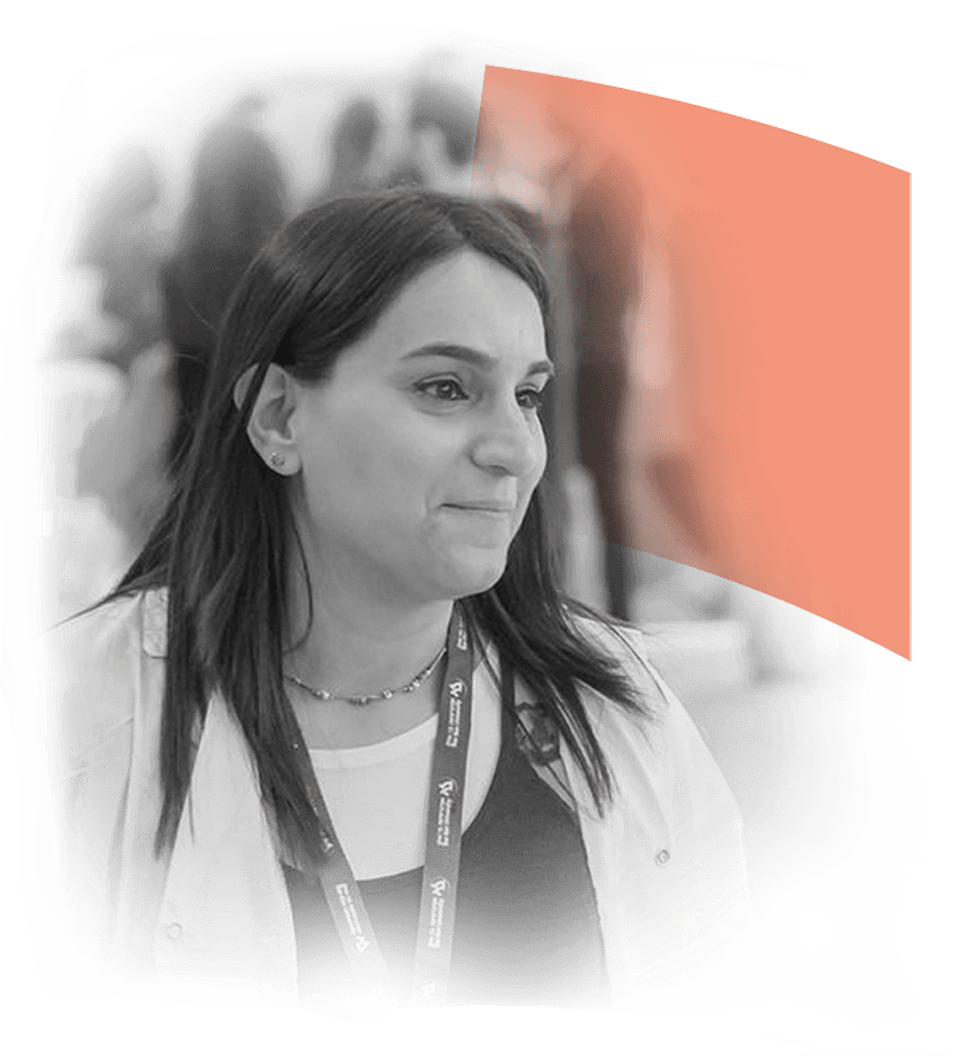 Smiling woman with dark hair wearing a lanyard and a light jacket, engaged in conversation at a professional event, with a vibrant orange graphic in the background.
