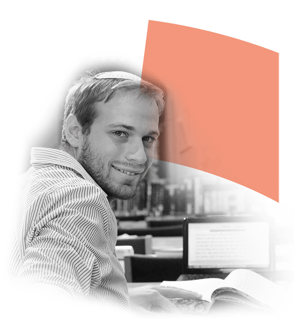 Smiling young man studying at a library with a laptop and an open book, wearing a striped shirt and a kippah, surrounded by bookshelves.