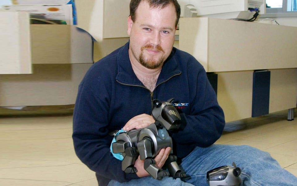 Man sitting on the floor with a robotic dog in his hands, showcasing the interaction between humans and technology in a modern setting. Another robotic dog is placed beside him, highlighting advancements in robotic pets.