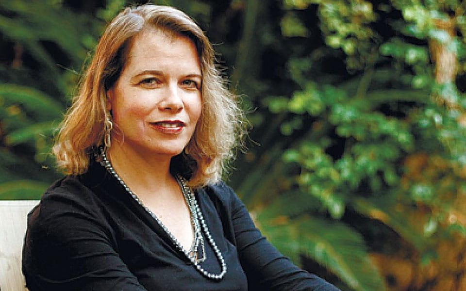 Portrait of a woman with shoulder-length blonde hair, wearing a black top and pearl necklace, seated outdoors with lush green foliage in the background.