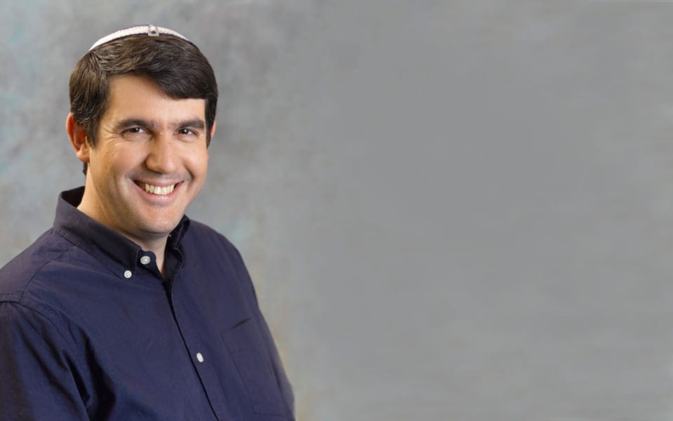 Smiling man wearing a kippah, dressed in a dark button-up shirt, posing against a softly blurred gray background.