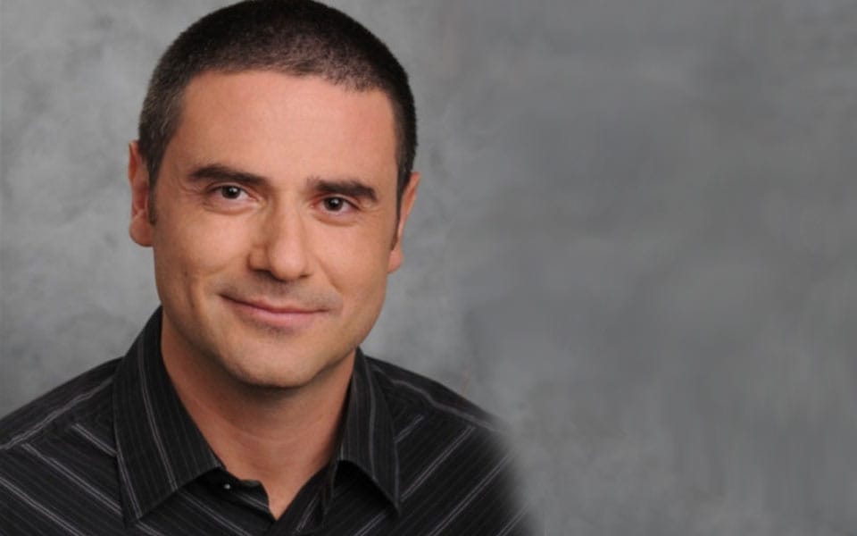 Professional headshot of a man with short hair and a slight smile, wearing a black striped shirt against a gray background. Ideal for business profiles or personal branding.
