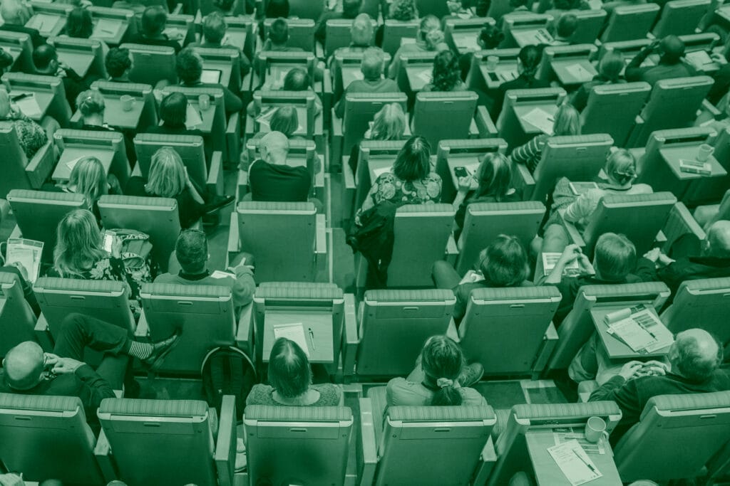 High-angle, monochromatic view of a conference room filled with attendees seated in rows of green chairs. Each chair has a small desk attached.