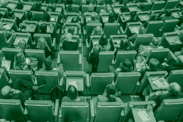 High-angle, monochromatic view of a conference room filled with attendees seated in rows of green chairs. Each chair has a small desk attached.