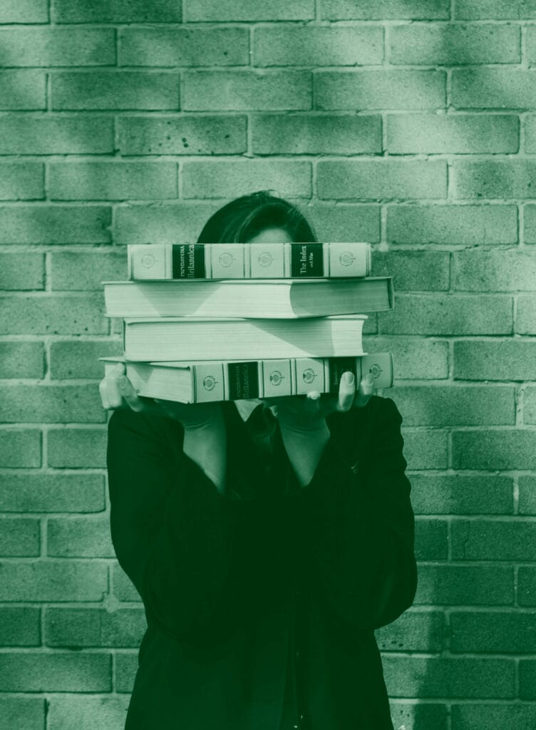 A person stands in front of a brick wall, their face hidden behind a stack of books. The books appear to be encyclopedias.