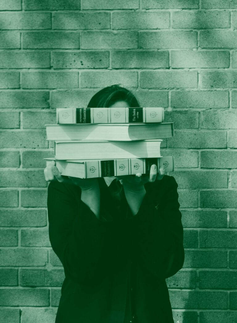 A person stands in front of a brick wall, their face hidden behind a stack of books. The books appear to be encyclopedias.