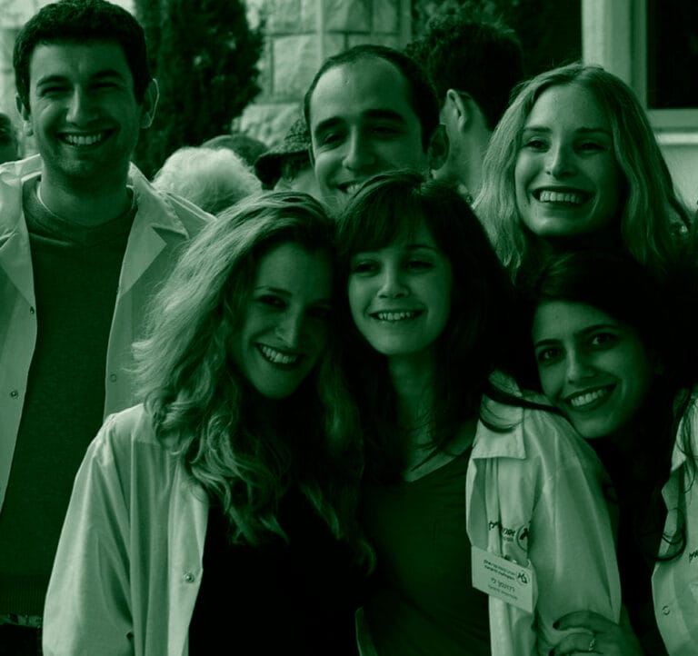 A group of young adults, mostly women, stand close together smiling for a photo. They are wearing white coats and are possibly at a medical or scientific event, as suggested by the name tags visible on some of the coats. The image is in sepia tone.
