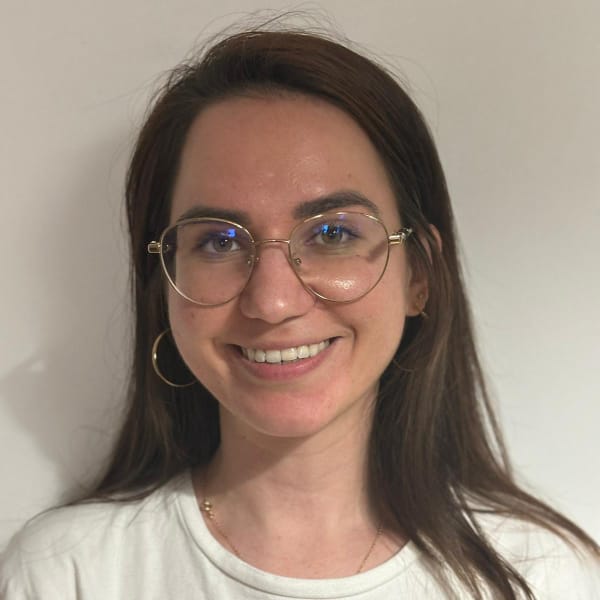 Close-up portrait of a young woman with long brown hair wearing gold-rimmed glasses and a white shirt.