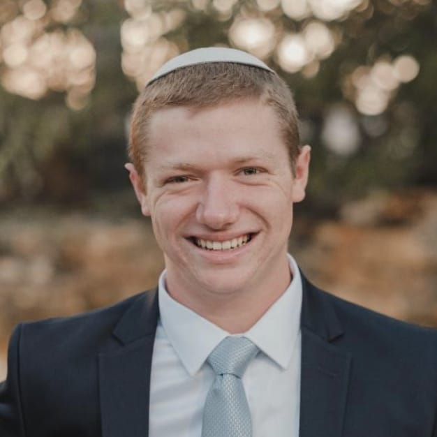 Close-up portrait of a young man wearing a kippah and a suit. He is smiling and looking directly at the camera. The background is blurred, suggesting an outdoor setting.