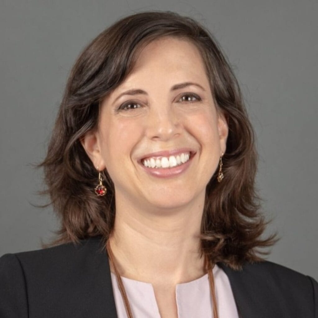Headshot of a smiling woman with shoulder-length brown hair wearing a black blazer and a light purple shirt.