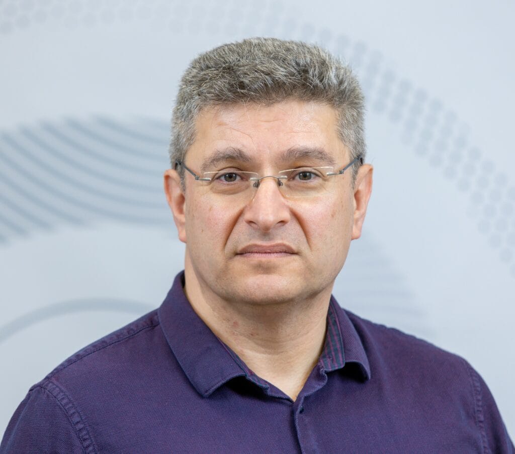 Close-up portrait of a man with short graying hair wearing glasses and a dark purple collared shirt.