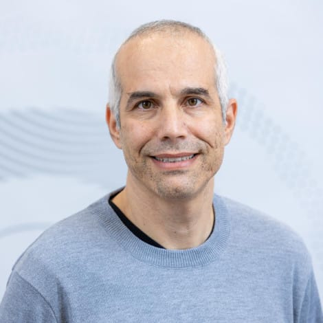 Close-up portrait of a man with short graying hair, wearing a gray sweater. He has a friendly expression. The background is a light gray with a subtle pattern.