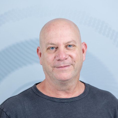 Close-up portrait of a bald, middle-aged man with blue eyes, wearing a dark gray t-shirt, against a light blue background with abstract patterns.