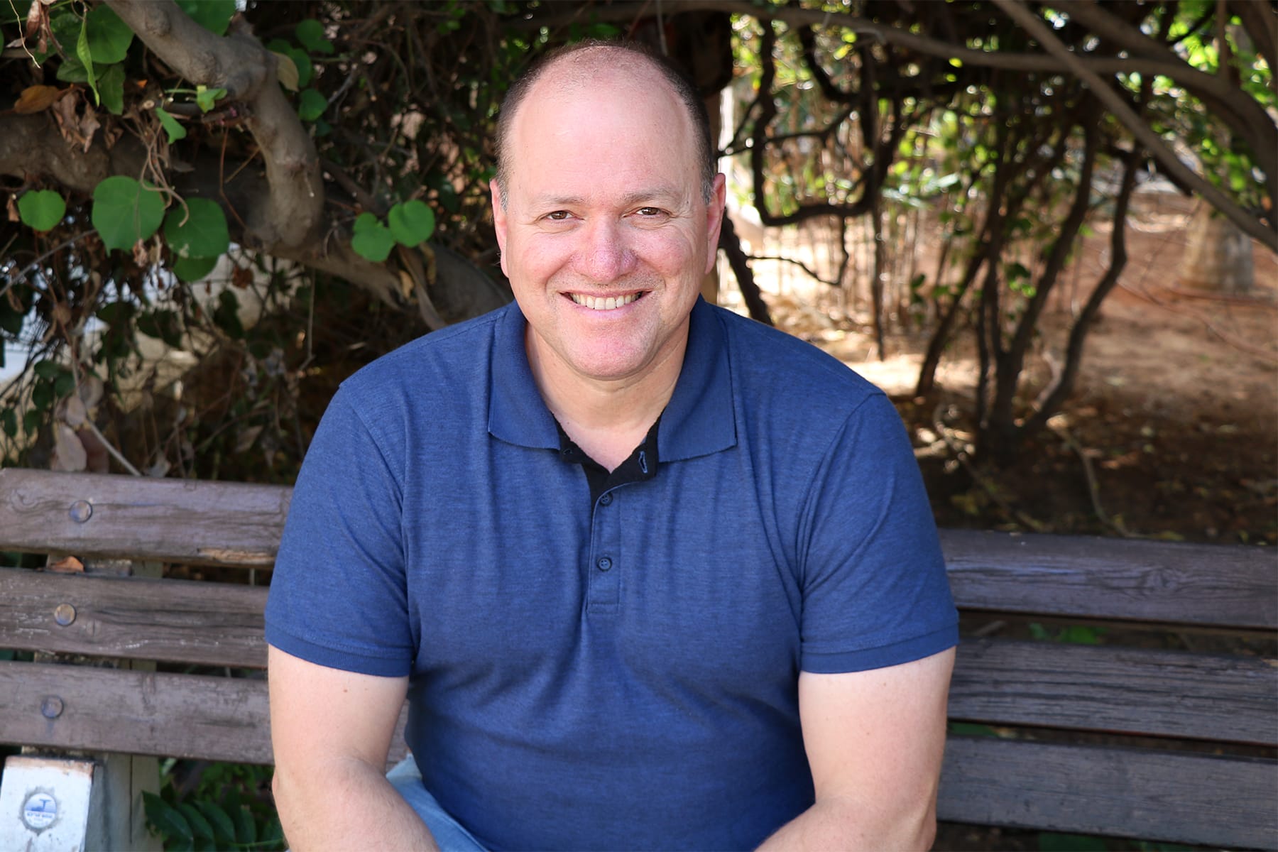 A bald, smiling man in a blue polo shirt sits on a wooden bench outdoors.